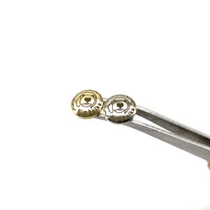 Op de afbeelding: Twee kleine, ronde metalen knopen met een honden gezicht ontwerp en het woord "Bimbulka" gegraveerd. Eén knoop is goudkleurig, de andere is zilverkleurig. De knopen worden vastgehouden door een zilveren pincet tegen een witte achtergrond.