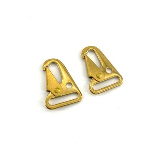 May include: Two brass-colored swivel snap hooks. Each hook has a triangular body with a spring-loaded clasp and a rectangular loop. The hooks are designed for attaching to straps or other items. The background is white.