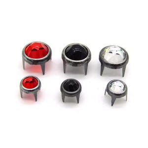 May include: Six metal studs with a vintage appearance. The studs have a dark gray base and three prongs. The tops are red, black, and clear, faceted, and jewel-like. The studs are arranged on a white background.