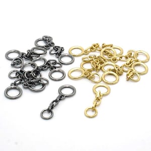 Könnte beinhalten: Eine Auswahl an Metallketten und Ringen in zwei Farben: Dunkelgrau und Gold. Die Glieder variieren in Größe und Stil, einige weisen kleine Verbindungsglieder auf, andere bestehen aus einfachen Ringen. Die Artikel sind auf einer weißen Oberfläche verstreut.