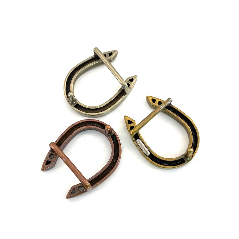 K&ouml;nnte beinhalten: Drei hufeisenf&ouml;rmige Metallschnallen in Bronze-, Silber- und Goldt&ouml;nen. Jede Schnalle hat ein D-f&ouml;rmiges Design mit einem Steg oben und einem dekorativen Element an jedem Ende. Die Schnallen befinden sich auf wei&szlig;em Hintergrund.