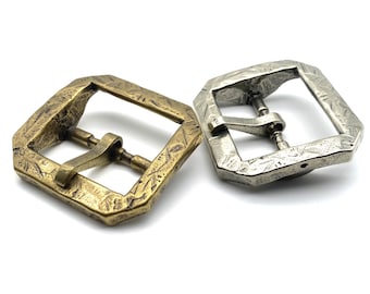 May include: Two metal belt buckles, one gold and one silver, with an ornate design. Both buckles have a square shape with rounded corners and a textured surface.