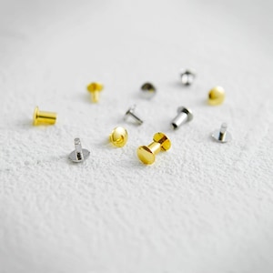 Könnte beinhalten: Eine Auswahl kleiner, metallischer Nieten in Gold- und Silbertönen. Die Nieten haben verschiedene Formen, darunter flache, runde und zylindrische Designs. Sie sind auf einer weißen Oberfläche verstreut und zeigen ihre reflektierenden Eigenschaften.