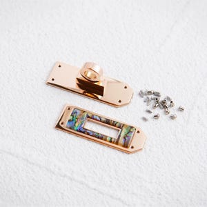 Puede incluir: Juego de herrajes metálicos para bolsos en tono oro rosa. Incluye un cierre rectangular con un anillo, una base rectangular con incrustaciones de abulón y pequeños tornillos plateados. La incrustación de abulón presenta iridiscencias azules, verdes y marrones.