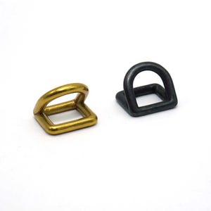 May include: Two metal D-rings. One is gold-colored, the other is dark gray. Each ring has a square base with a rounded top loop. The gold ring is on the left, and the dark gray ring is on the right, both against a white background.