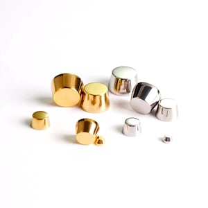 May include: A collection of metallic thimble-shaped objects in gold and silver tones. The objects vary in size, with a flat top and a conical body. The reflective surfaces create highlights and shadows against the white background.