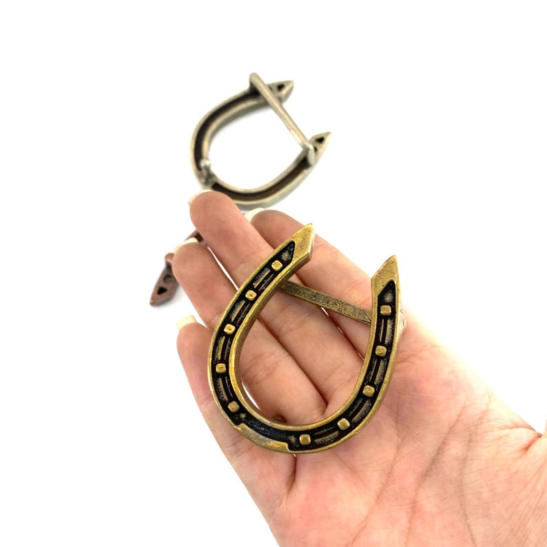 K&ouml;nnte beinhalten: Zwei hufeisenf&ouml;rmige G&uuml;rtelschnallen aus Metall. Eine wird in einer Hand gehalten und zeigt ein detailliertes Design mit goldener und schwarzer Oberfl&auml;che. Die andere Schnalle ist silberfarben. Die Schnallen sind wahrscheinlich f&uuml;r G&uuml;rtel oder Accessoires im Western-Stil.