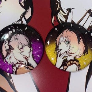 May include: Two round, colorful buttons with images of anime characters. The button on the left has a purple and black background with a white character with pink hair. The button on the right has a gold and black background with a white character with pink hair.