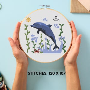 May include: A cross-stitch pattern of a blue dolphin jumping out of the water with green seaweed and a red crab. The pattern is framed in a wooden hoop and the text "STITCHES: 120 X 107" is printed below the hoop.
