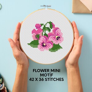 May include: A close-up of a cross-stitch embroidery hoop with a pink flower design. The hoop is held in a person's hands. The text on the hoop reads "FLOWER MINI MOTIF 42 X 36 STITCHES".