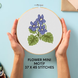 May include: A cross-stitch pattern of a purple flower with green leaves in a wooden embroidery hoop. The text below the hoop reads "FLOWER MINI MOTIF 37 X 45 STITCHES".