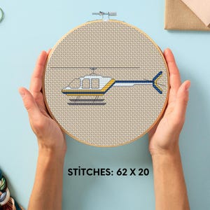 Pode incluir: Padrão de ponto cruz de um helicóptero branco e amarelo com detalhes azuis em um tecido bege. O padrão está em um aro de bordado redondo e o texto "STITCHES: 62 X 20" está abaixo do aro.