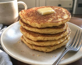 Op de afbeelding: Een stapel van vijf goudbruine pannenkoeken op een gespikkeld gebroken wit keramieken bord. Een vierkant blokje gele boter ligt op de bovenste pannenkoek. Een zilveren metalen vork ligt aan de rechterkant van het bord en een mok is zichtbaar.