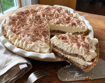 Op de afbeelding: Een ronde tiramisu-taart in een witte, gegolfde keramische schaal, met lagen in koffie gedoopte lange vingers en mascarponecrème, afgewerkt met een dikke laag slagroom en cacaopoeder, waarbij één punt wordt opgetild met een metalen taartschep.