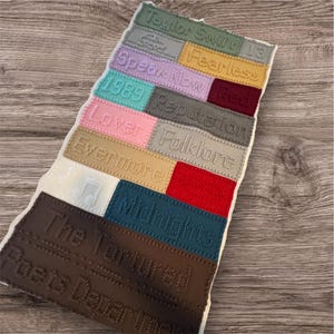 May include: A colourful knitted wall hanging featuring the names of Taylor Swift's albums, including "Taylor Swift 13", "Fearless", "Speak Now", "1989", "Reputation", "Lover", "Folklore", "Evermore", "Midnights", "The Tortured", and "Posts Departme".