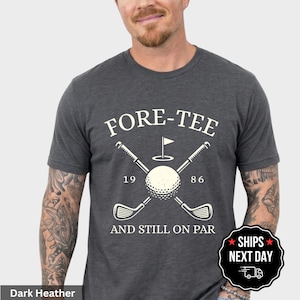 May include: A dark heather gray short-sleeve t-shirt featuring a white graphic of two crossed golf clubs, a golf ball, and a flag. The text reads FORE-TEE, 19 86, and AND STILL ON PAR. The design is centered on the chest of the crew neck garment.