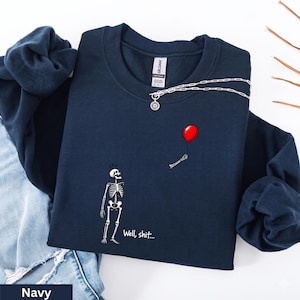 May include: A navy blue crewneck sweatshirt featuring a graphic of a white skeleton looking up at a floating red balloon held by a detached skeletal hand. The text "Well, shit..." is printed in white below the skeleton on the front of the garment.