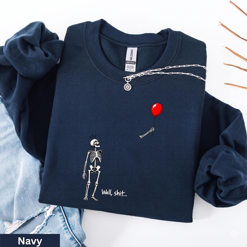May include: A navy blue crewneck sweatshirt featuring a graphic of a white skeleton looking up at a floating red balloon held by a detached skeletal hand. The text "Well, shit..." is printed in white below the skeleton on the front of the garment.