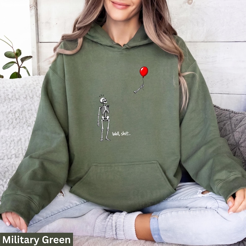 May include: A military green hooded sweatshirt featuring a graphic of a skeleton looking up at a red balloon floating away, with a skeletal hand still holding the string. The text "Well, shit..." is printed in white below the skeleton figure.
