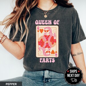 A woman wearing a black t-shirt with a graphic design of a woman holding a cigarette and the text "Queen of Farts" printed on it.