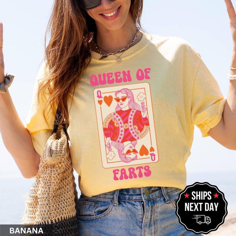 A woman wearing a yellow t-shirt with a graphic design of a woman holding a deck of playing cards, specifically a queen of hearts. She is smiling and holding her hands up.