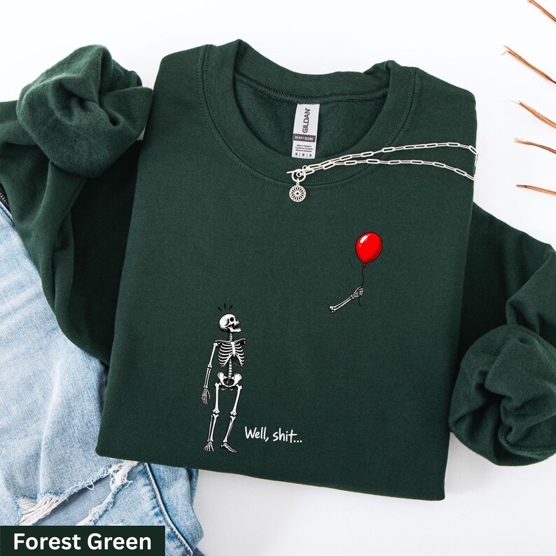 May include: A forest green crewneck sweatshirt featuring a graphic of a skeleton looking up at a floating red balloon held by a disembodied skeletal hand. The text "Well, shit..." is printed in white below the skeleton. A silver chain rests on the collar.