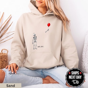 May include: A sand-colored hooded sweatshirt featuring a graphic of a skeleton looking up at a detached skeletal hand holding a red balloon. The text "Well, shit..." is printed in black below the skeleton on the front of the garment.