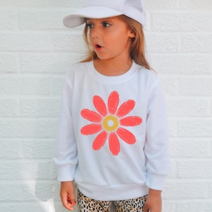 A young girl wearing a white hat and a white sweatshirt with a large red flower design stands against a white brick wall.
