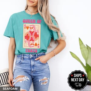 A woman wearing a teal t-shirt with a graphic design of a woman playing cards, specifically a queen of hearts, and blue jeans.