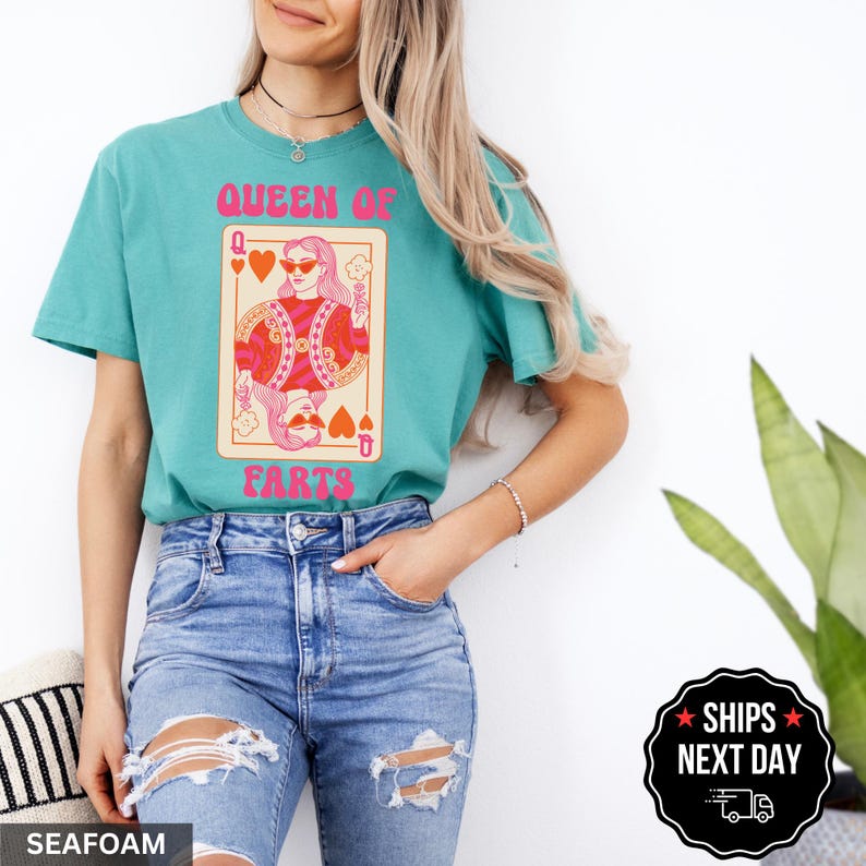 A woman wearing a teal t-shirt with a graphic design of a woman playing cards, specifically a queen of hearts, and blue jeans.