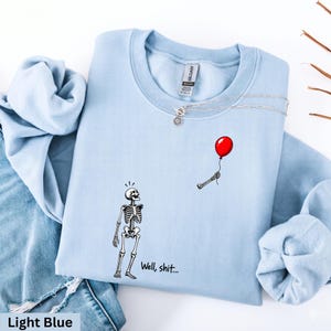 May include: A light blue crewneck sweatshirt featuring a graphic of a skeleton looking up at a floating red balloon held by a detached skeletal hand. The text "Well, shit..." is printed in black below the skeleton on the front of the garment.