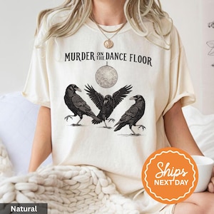 May include: Cream-colored t-shirt featuring the text "Murder on the Dance Floor" above a graphic of three black crows and a disco ball. The shirt has short sleeves and a crew neck.