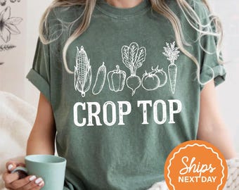 May include: A moss green crop top featuring a white graphic of various vegetables, including corn, a cucumber, a pepper, a beet, a tomato, and a carrot. The words "CROP TOP" are printed in bold white letters below the vegetables.