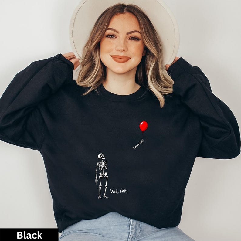 May include: A black long-sleeve crewneck sweatshirt featuring a graphic print of a white skeleton looking up at a red balloon held by a detached skeletal hand. The text "Well, shit..." is printed in white below the skeleton on the front.