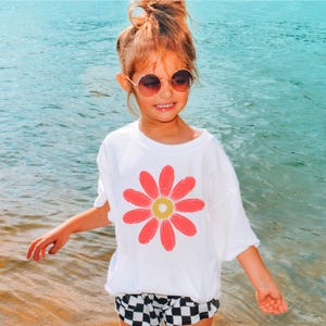 A young girl wearing a white t-shirt with a flower design and sunglasses stands near the water, smiling and enjoying the beach.