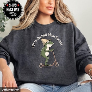 May include: Dark heather gray sweatshirt featuring a cartoon frog riding a scooter and wearing a hat. The text "Off to Commit Mom Foolery" is arched around the frog. The sweatshirt is a comfortable, casual style.