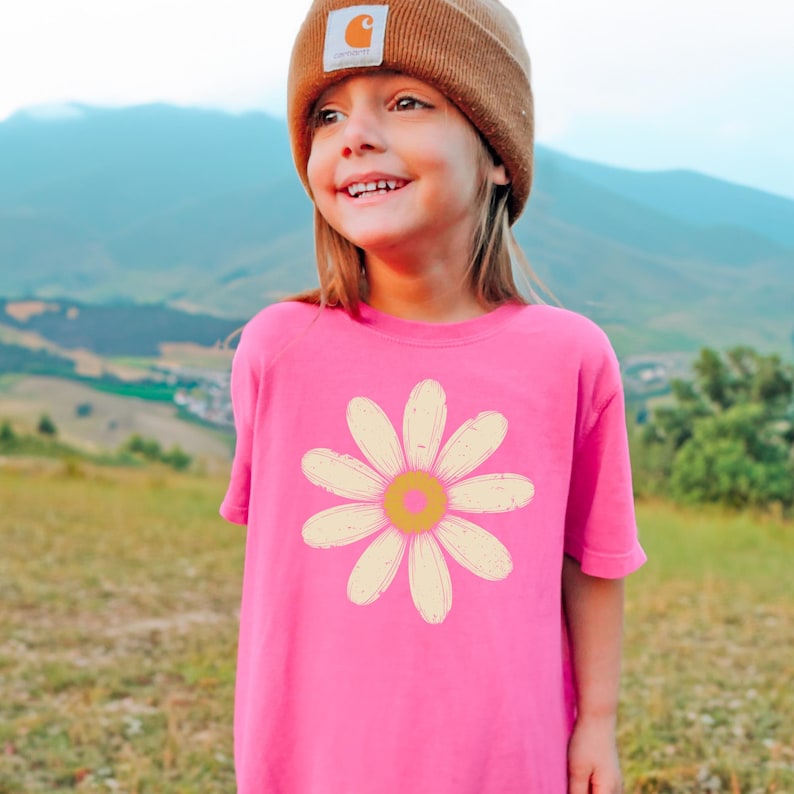 A young girl wearing a pink t-shirt with a flower design and a brown beanie stands in a grassy field with mountains in the background.