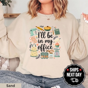 May include: A sand-colored crewneck sweatshirt featuring a graphic print with the text I'll be in my office surrounded by illustrations of a stand mixer, rolling pin, pie, whisk, cookbook, oven mitt, apron, piping bag, cupcake, eggs, and scale.