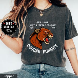 May include: Dark gray t-shirt with a cougar graphic and the text "STILL HOT JUST A LITTLE FLASHY EST 2025 COUGAR PUBERTY." The shirt has a vintage look.