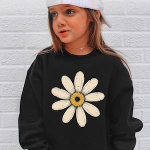 A young girl wearing a black sweatshirt with a large white flower design on it, standing against a white brick wall.