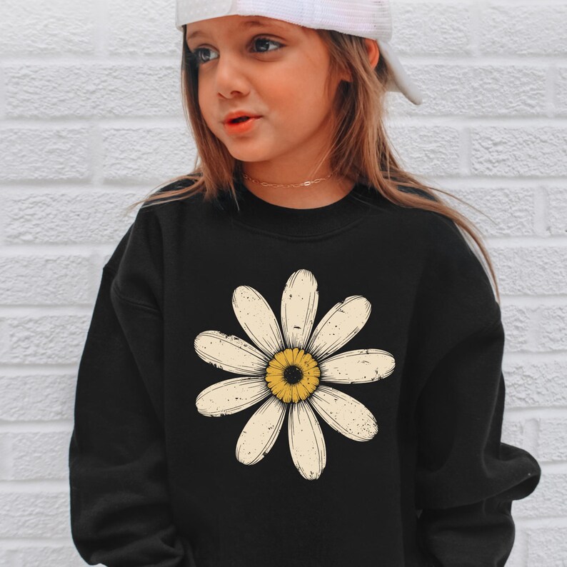A young girl wearing a black sweatshirt with a large white flower design on it, standing against a white brick wall.