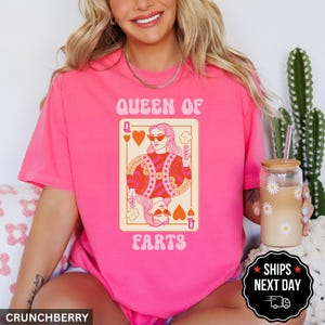 A woman wearing a pink t-shirt with a graphic of a woman holding a deck of cards and the text "Queen of Farts" printed on it. She is sitting next to a cactus plant and holding a drink.
