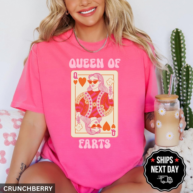 A woman wearing a pink t-shirt with a graphic of a woman holding a deck of cards and the text "Queen of Farts" printed on it. She is sitting next to a cactus plant and holding a drink.