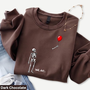 May include: A dark chocolate brown crewneck sweatshirt featuring a graphic of a skeleton looking up at a floating red balloon held by a disembodied skeletal hand, with the text "Well, shit..." printed in white below the skeleton.
