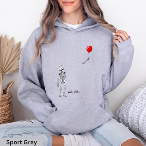 May include: A sport grey hooded sweatshirt featuring a graphic of a skeleton looking up at a red balloon held by a detached skeletal hand. The text "Well, shit..." is printed in black below the skeleton. The hoodie has long sleeves and a front pocket.