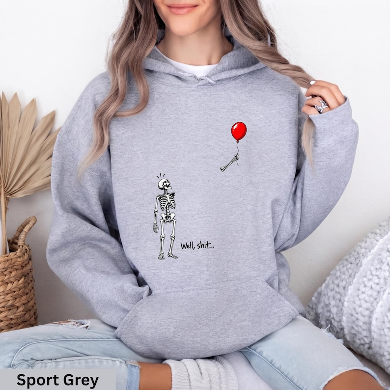 May include: A sport grey hooded sweatshirt featuring a graphic of a skeleton looking up at a red balloon held by a detached skeletal hand. The text "Well, shit..." is printed in black below the skeleton. The hoodie has long sleeves and a front pocket.