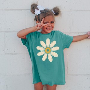 A young girl with pigtails is smiling and making a peace sign with her fingers, wearing a green t-shirt with a yellow flower design.