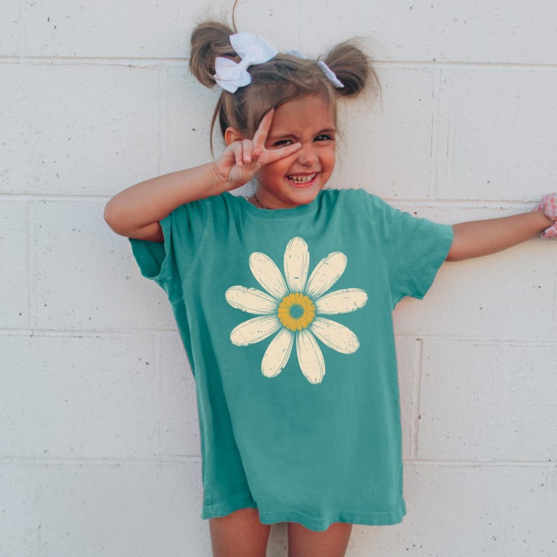 A young girl with pigtails is smiling and making a peace sign with her fingers, wearing a green t-shirt with a yellow flower design.