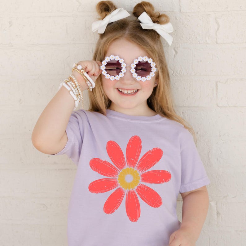 A young girl wearing a purple t-shirt with a flower design, sunglasses, and a bow in her hair is smiling and posing for the camera.