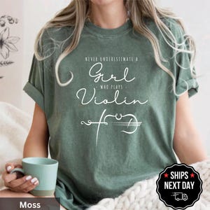 May include: Moss green t-shirt with the text "Never Underestimate a Girl Who Plays Violin". The design features a violin illustration. The shirt is a comfortable, casual wear item.